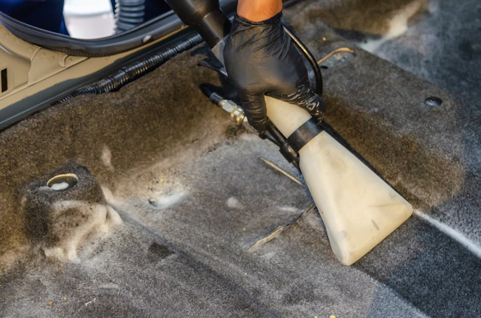 Close-up view of a carpet cleaning vacuum cleaner with a clear water tank filled with soapy water, connected to a flexible, patterned hose. The appliance is positioned on a beige carpeted floor in a r